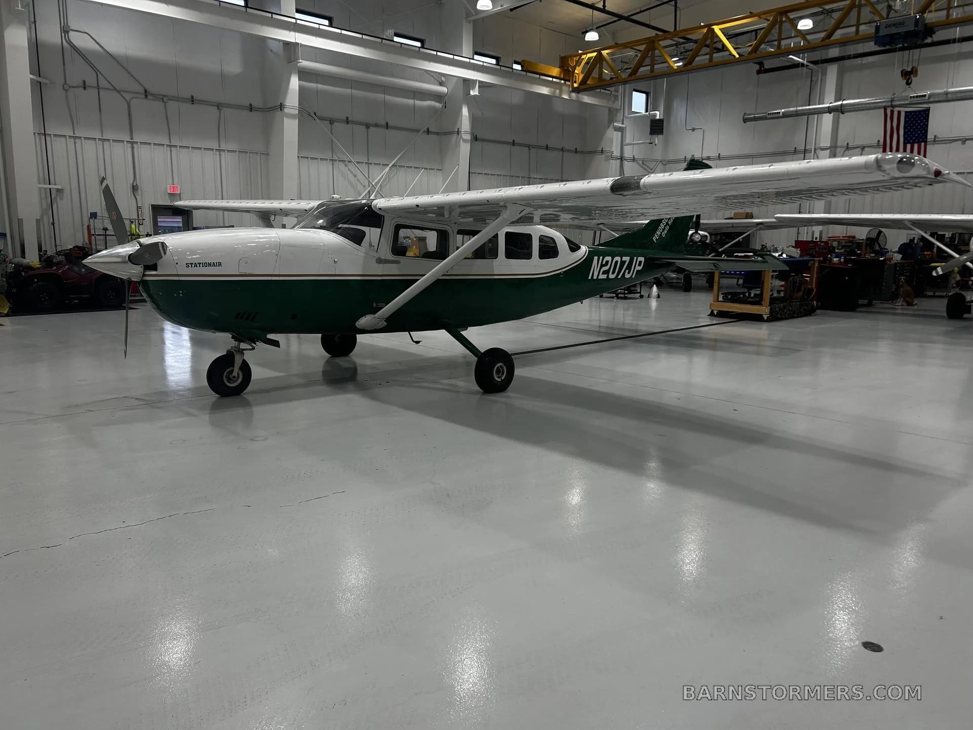 A white and green Cessna 207 Stationair, registration N207JP, is parked in a hangar, viewed from the front-right side.