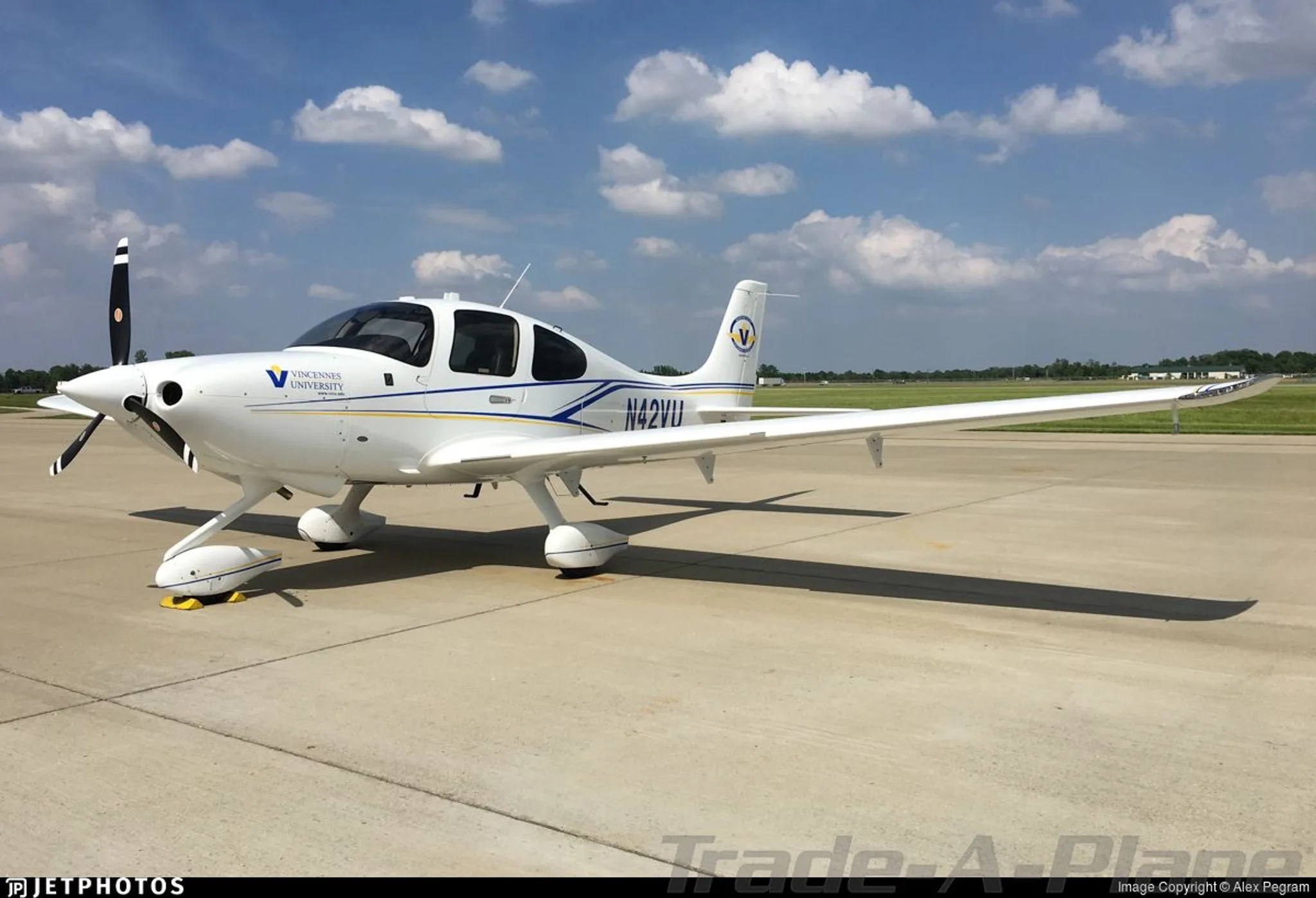 A white single-engine propeller aircraft, N42VU, with blue and yellow stripes and Vincennes University logo, parked on an airfield under a blue sky with scattered clouds.
