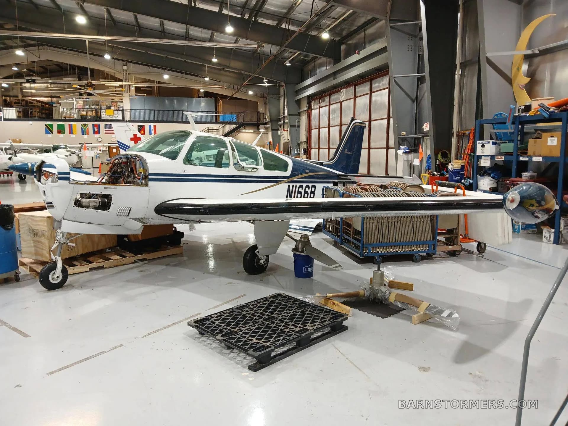 A white and blue Beechcraft Bonanza (N166B) with its engine cowling removed, parked in a hangar.