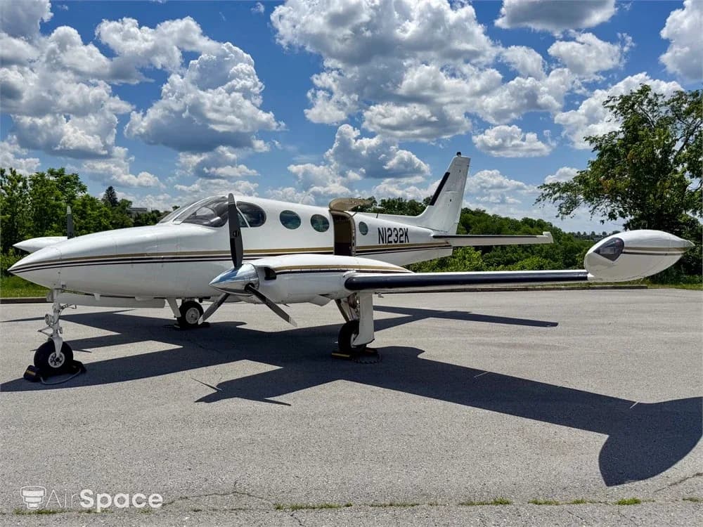 A white twin-engine aircraft with gold and brown stripes parked on an asphalt ramp under a partly cloudy sky.
