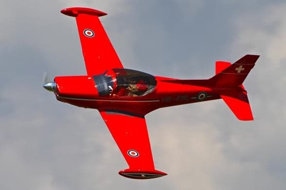 A red aircraft, HB-EML, in flight against a green hillside.
