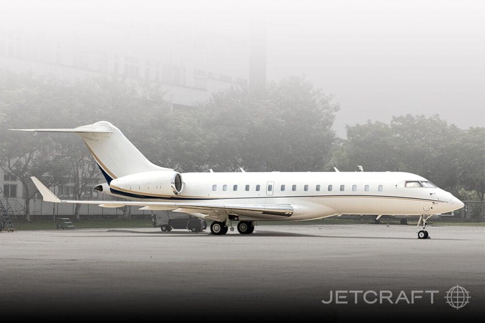 A white Bombardier Global Express business jet parked on the tarmac in foggy conditions.