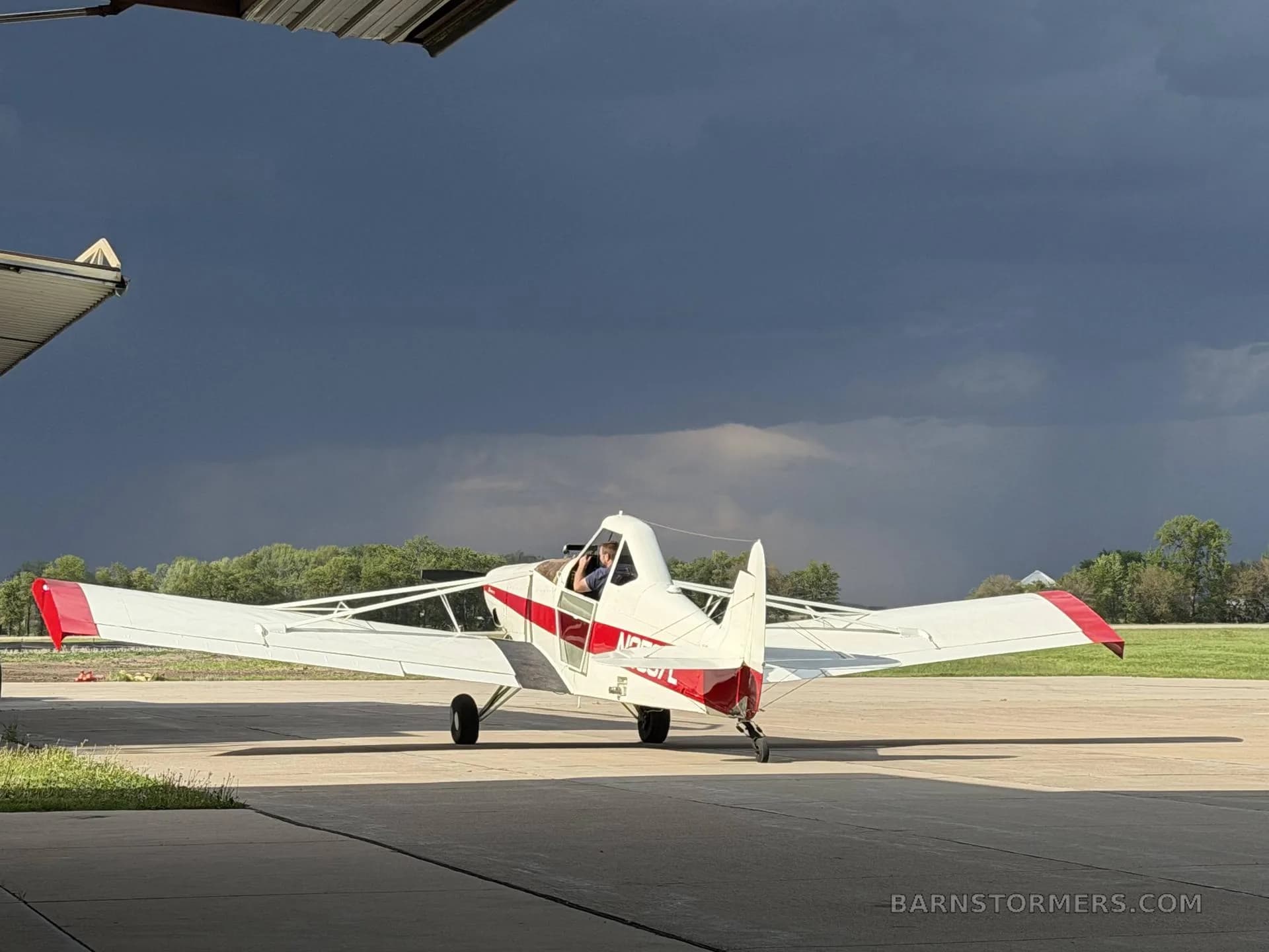 A white and red agricultural aircraft, seen from the rear-left, with a pilot in the cockpit, parked on an airfield under a dark, cloudy sky.