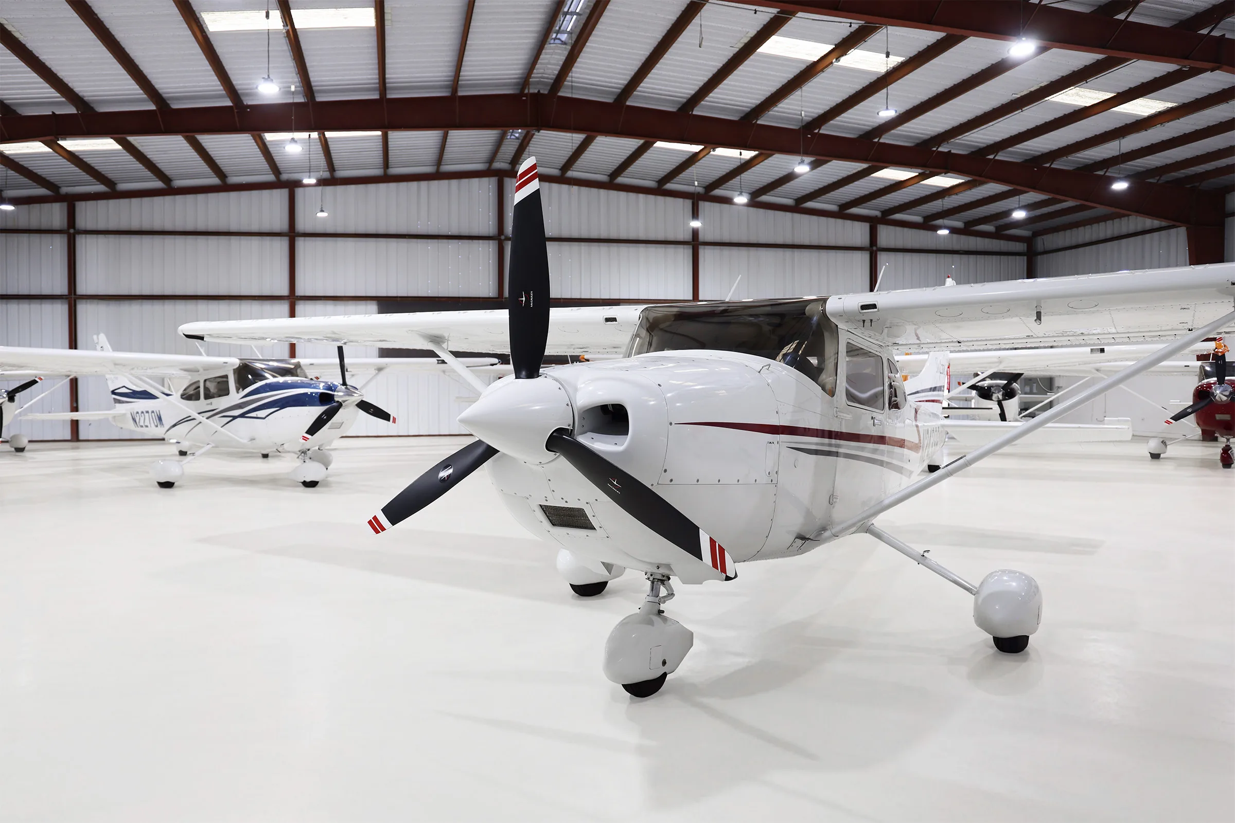 A white Cessna 182 Skylane with red and grey stripes, parked in a hangar, viewed from the front.