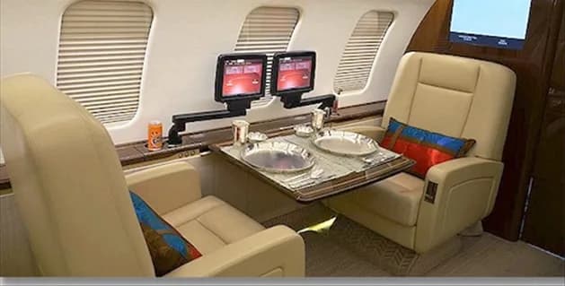 A luxurious aircraft interior with two beige leather seats facing each other, a dining table set for two, and dual entertainment screens mounted on an arm.