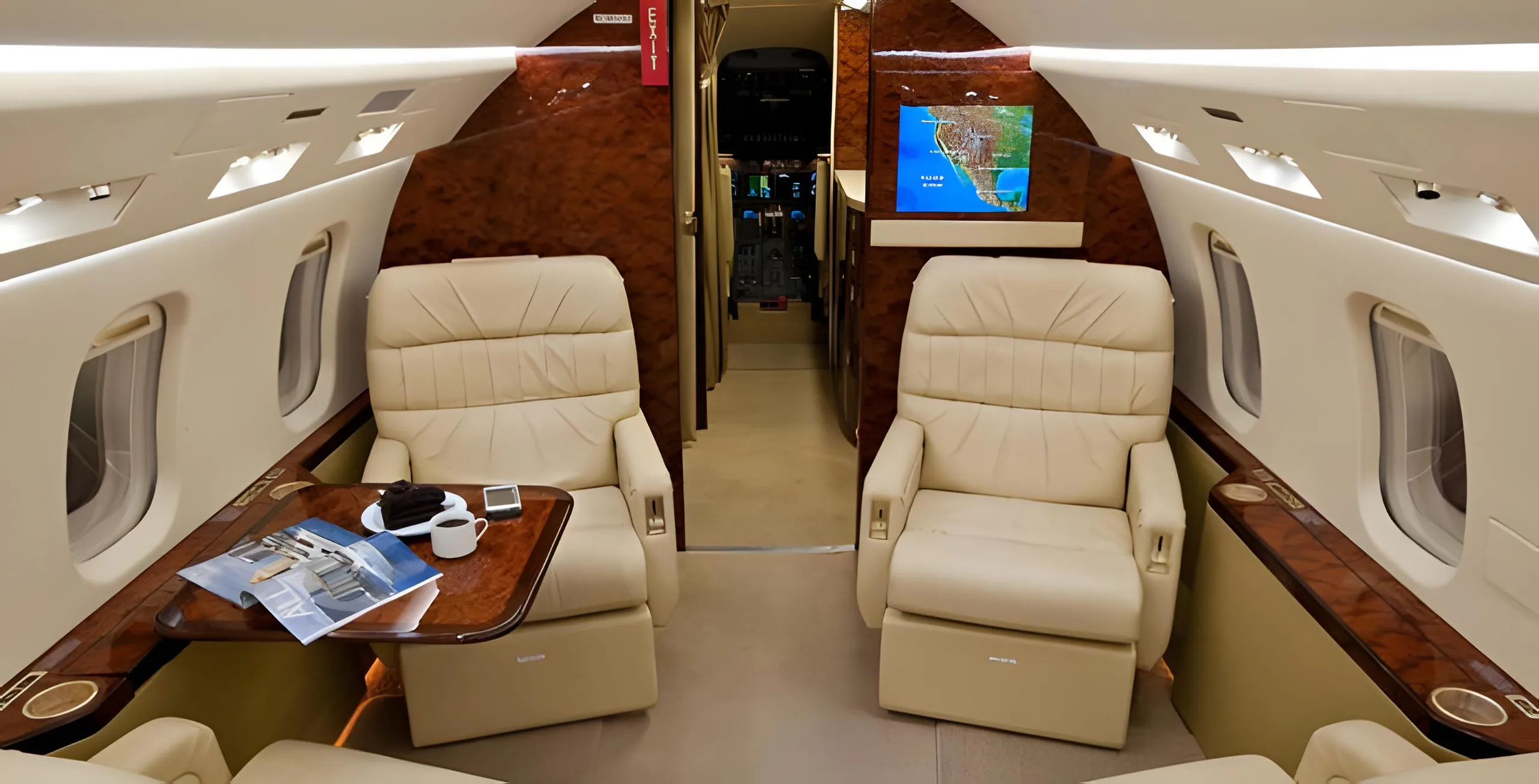 The aircraft's cabin interior looking forward, showing two beige leather seats, a wooden table with coffee and a magazine, and a map on a screen.