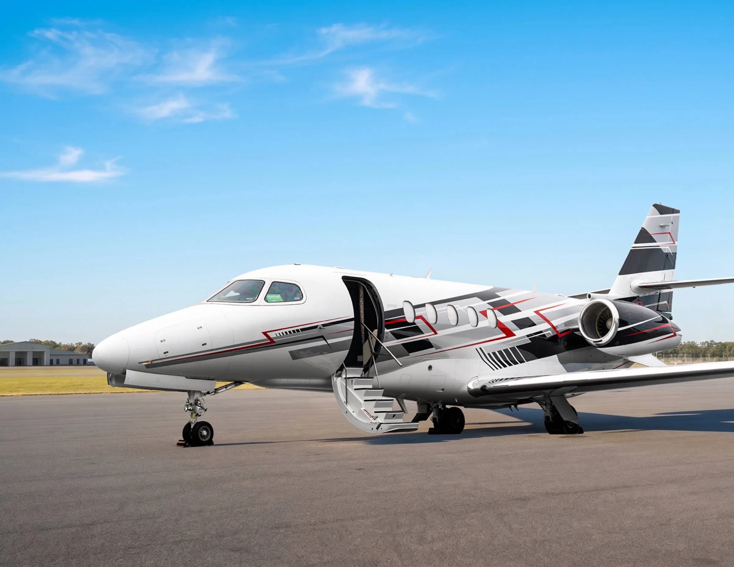 A front-side view of a white private jet with black and red geometric livery, parked with its main door open and stairs extended.