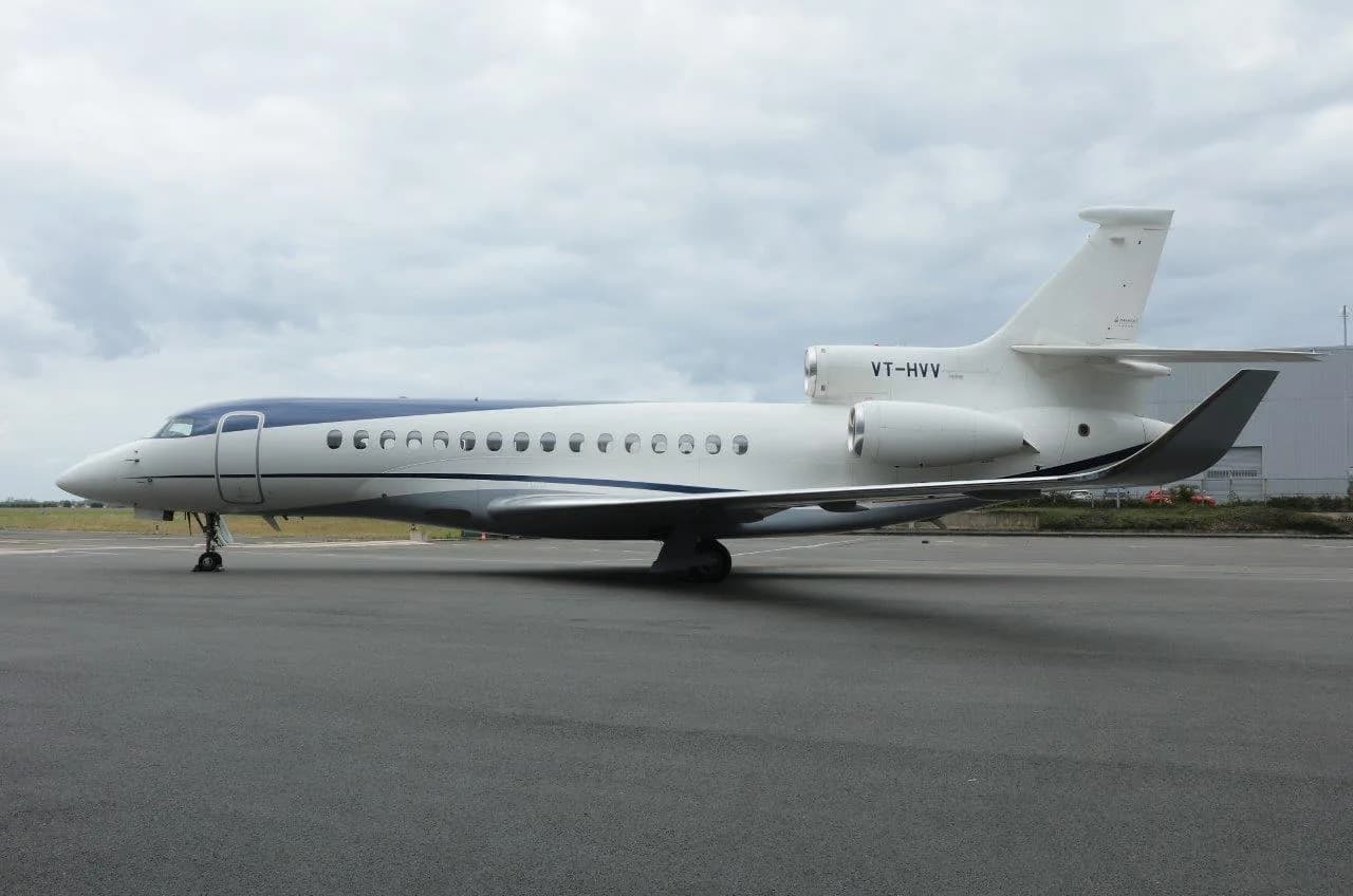A white and blue Dassault Falcon 900 business jet with registration VT-HVV parked on an airfield under a cloudy sky.