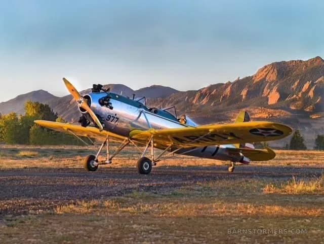 A silver and yellow biplane with '977' on the side, parked on a dirt runway with mountains in the background at sunset.