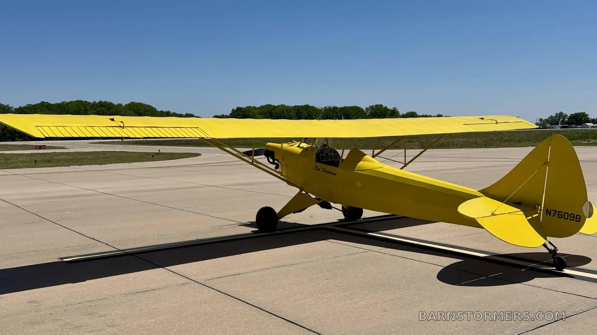 A bright yellow biplane, seen from the rear-right, parked on an asphalt tarmac with trees in the background.