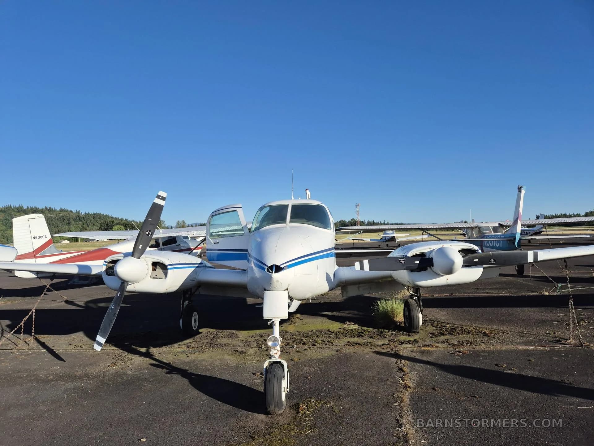 A white twin-engine aircraft with blue stripes, seen from the front with its door open, parked on an asphalt ramp under a clear blue sky.