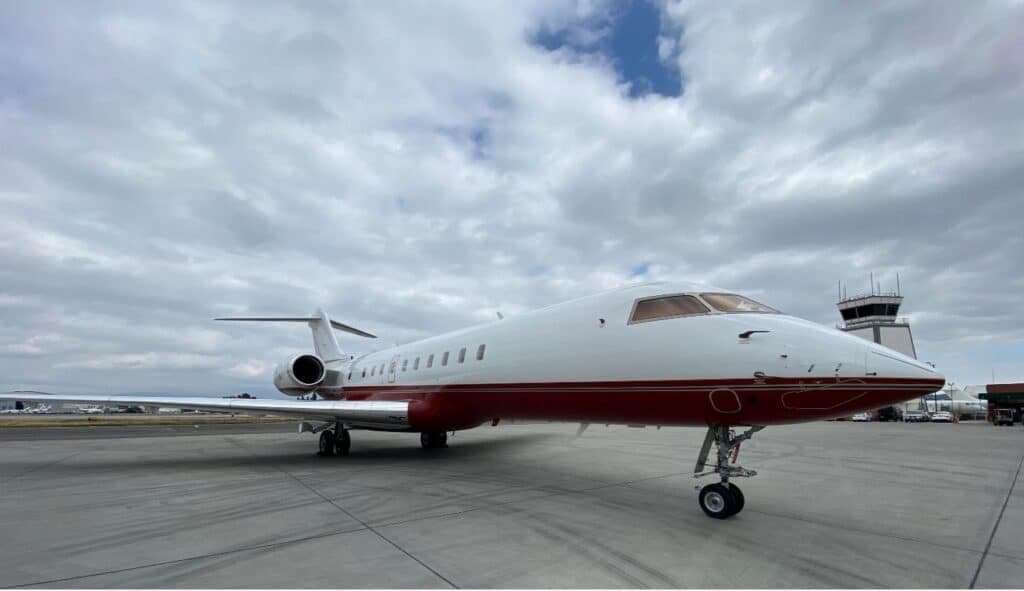 A white and red Bombardier Challenger 604 business jet parked on an asphalt tarmac with an air traffic control tower in the background.