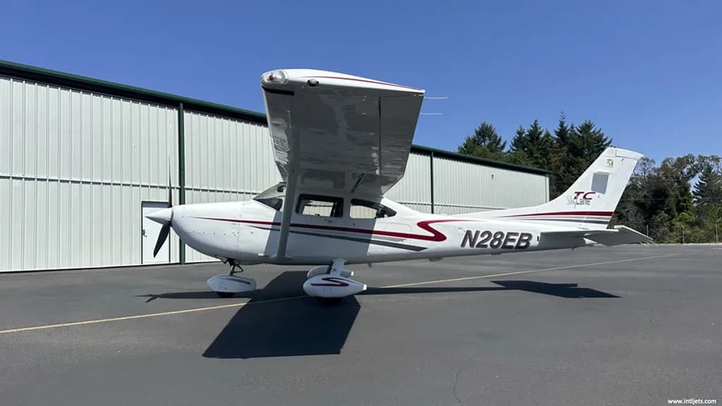 A white Cessna 182 Skylane with red and gray stripes, registration N28EB, parked on an asphalt ramp in front of a hangar.
