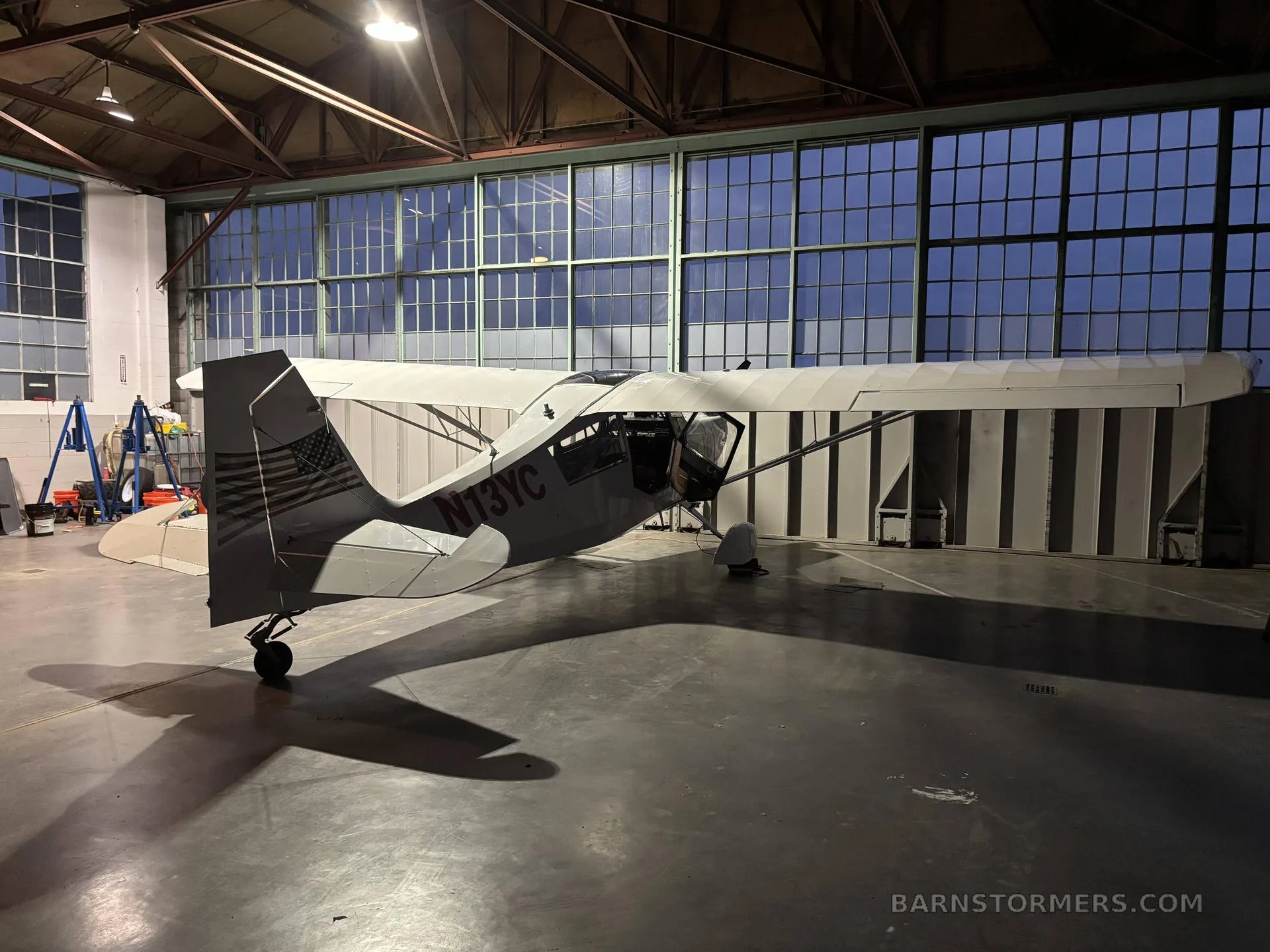 A light gray single-engine aircraft with N13YC registration and an American flag on the tail, viewed from the rear in a hangar.