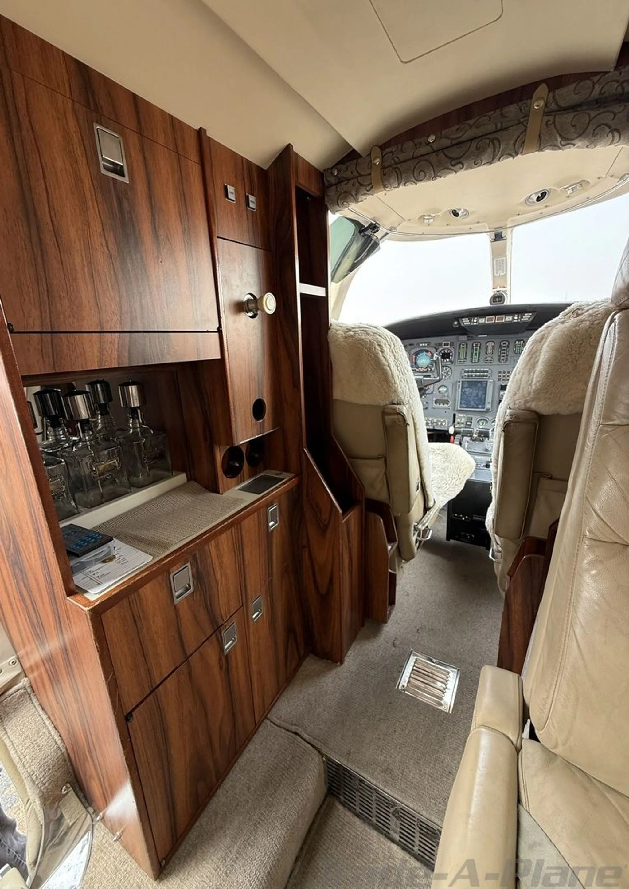 The interior cabin of the jet, showing wood cabinetry, a small bar area, and the cockpit beyond.