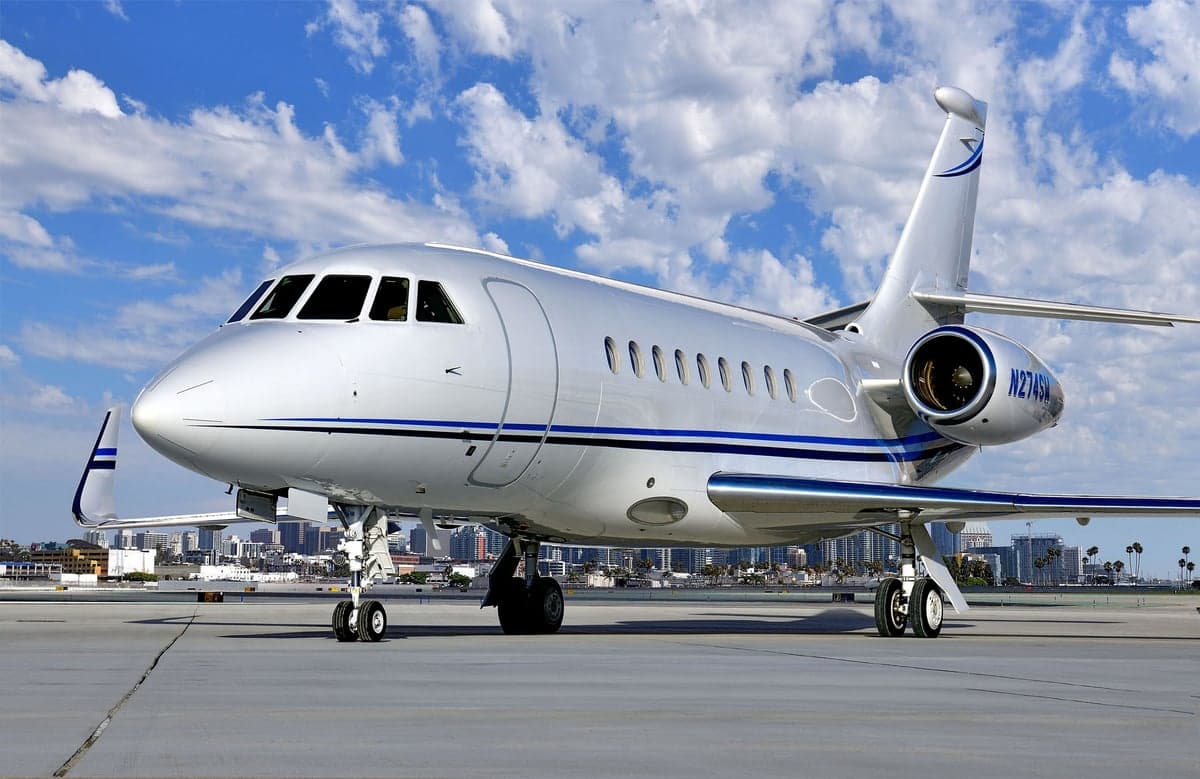 A Falcon aircraft sits on the tarmac with a city skyline in the background.