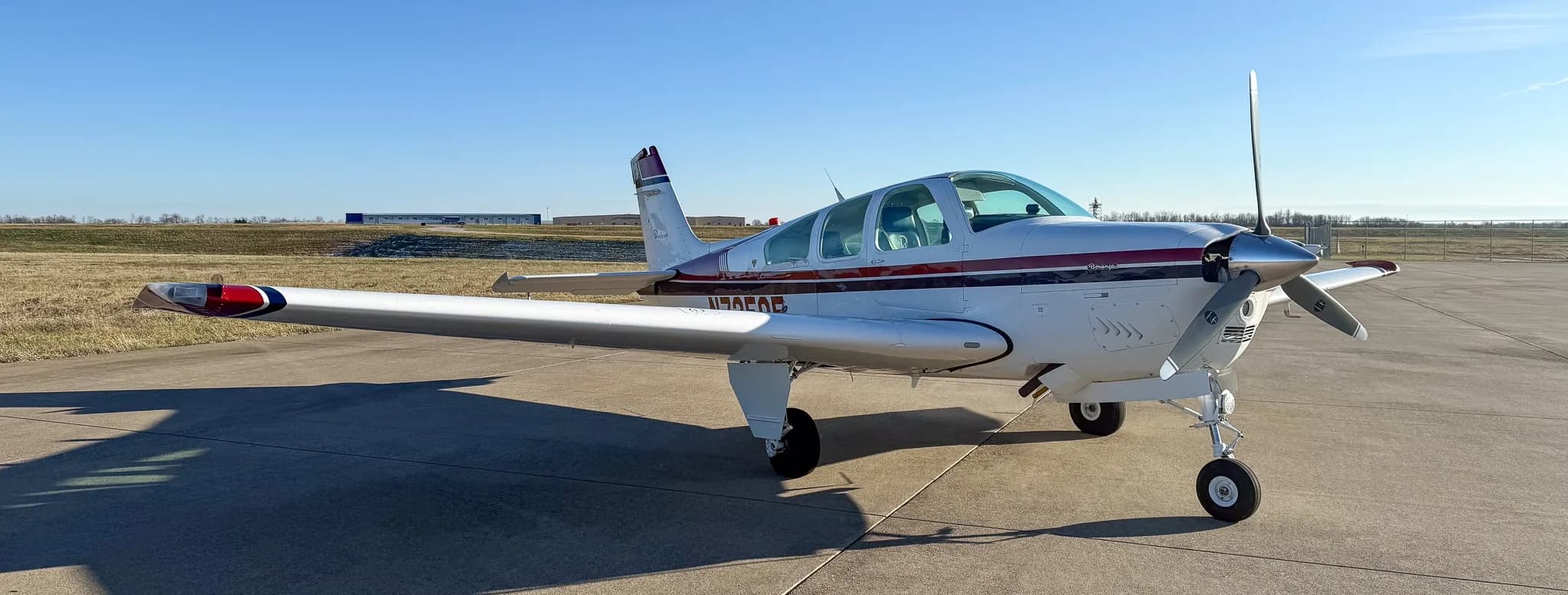 Front-left view of a white Beechcraft Bonanza with red and blue stripes on an airfield.
