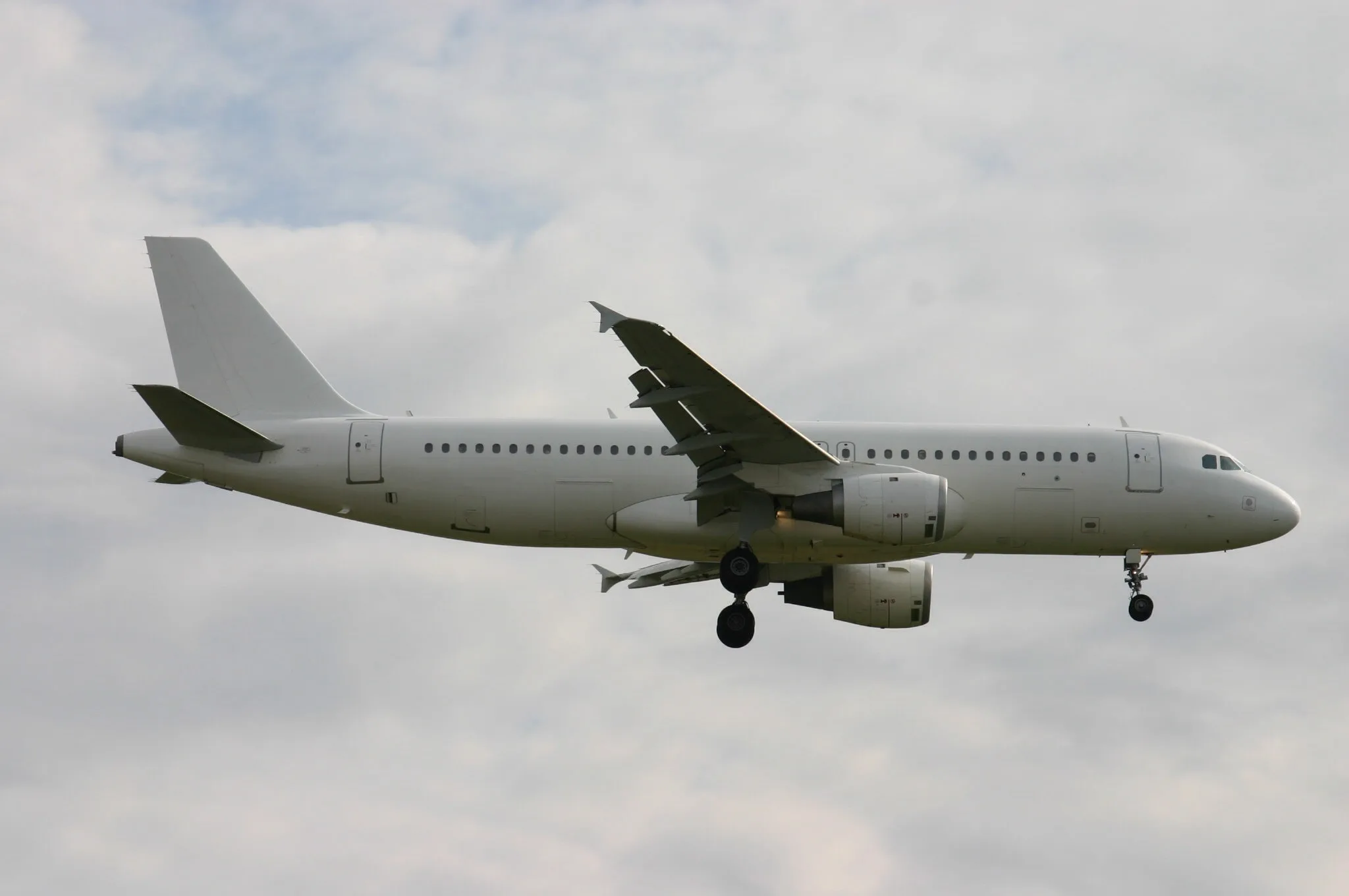A plain white Airbus A320 narrow-body jet with landing gear extended descends against a cloudy sky.