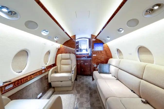 The spacious interior cabin of a private jet, featuring light-colored leather seating, a couch, and polished wood accents.