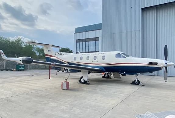 A white and dark blue Pilatus PC-12 turboprop aircraft with a red and gold stripe parked on a tarmac next to a hangar.