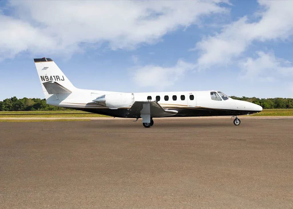 A white and black Cessna Citation business jet N941RJ with gold stripes is parked on a tarmac under a partly cloudy sky, viewed from the side.