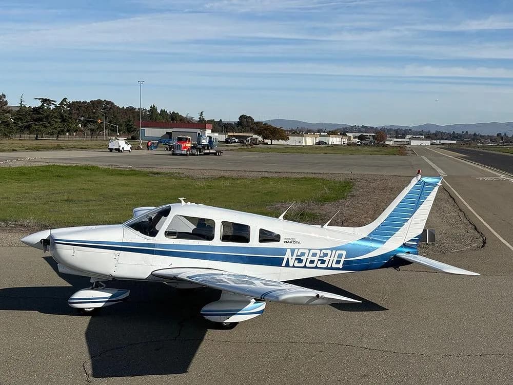 A white Piper Dakota aircraft with blue stripes, registration N3831Q, parked on an airfield with a runway in the background.