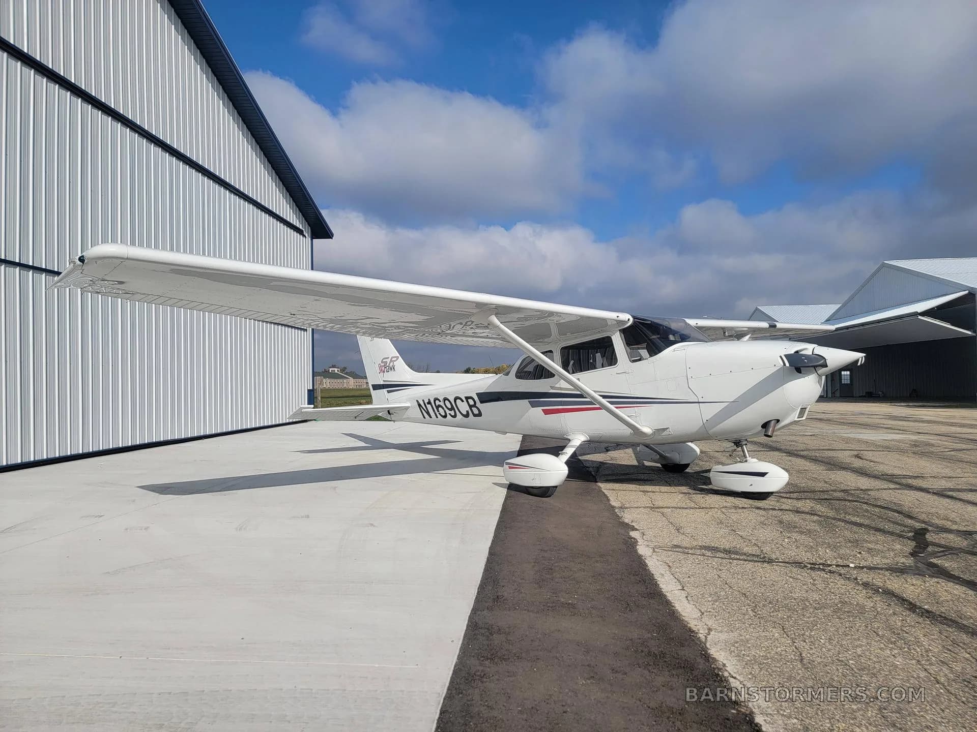 A white Cessna Skyhawk N169CB with black and red stripes is parked on an apron next to a white hangar under a partly cloudy sky.
