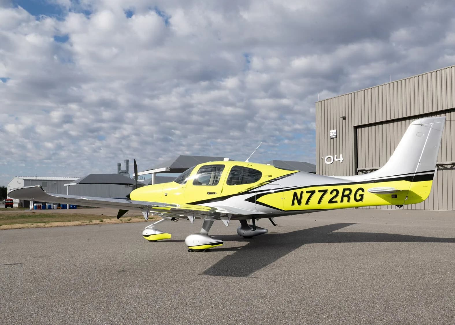A rear three-quarter view of the yellow and silver Cirrus SR22, N772RG, parked on an airfield with a hangar in the background.