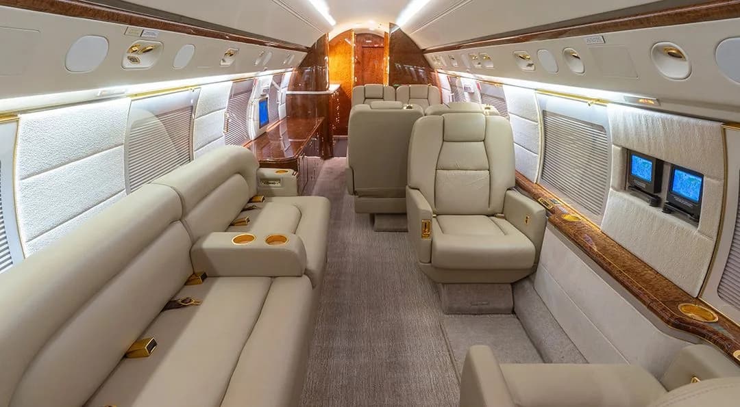 A view of the business jet's cabin, showing a beige leather sofa and individual seats with wood trim.