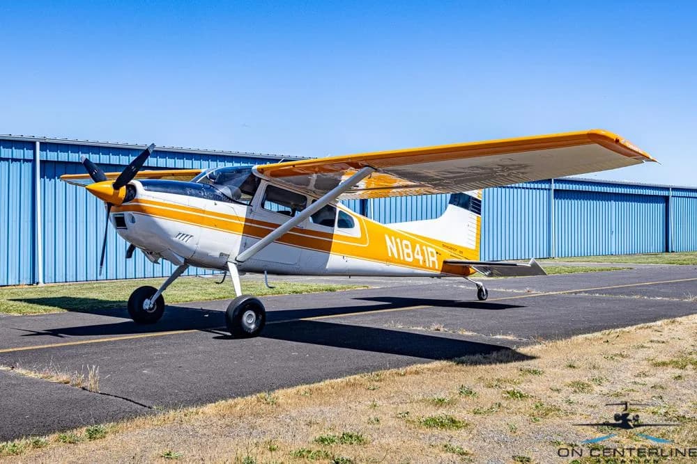 A white and orange Cessna 185 Skywagon (N1841R) parked on an asphalt ramp with blue hangars in the background.