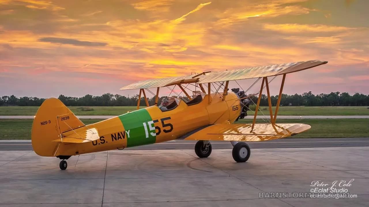 A yellow biplane with U.S. Navy markings and the number 155 on its fuselage, parked on a runway at sunset.