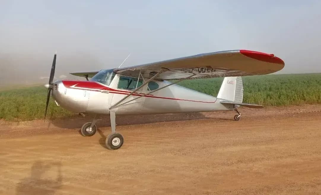 A silver and red Cessna 170 aircraft is parked on a dirt field.