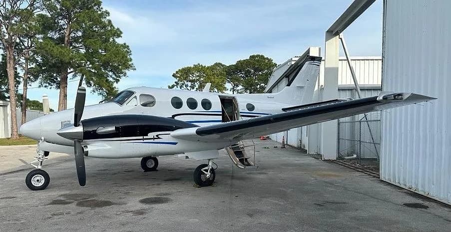 A white and black Beechcraft King Air with blue accent stripes parked on an asphalt apron with its airstair door open.
