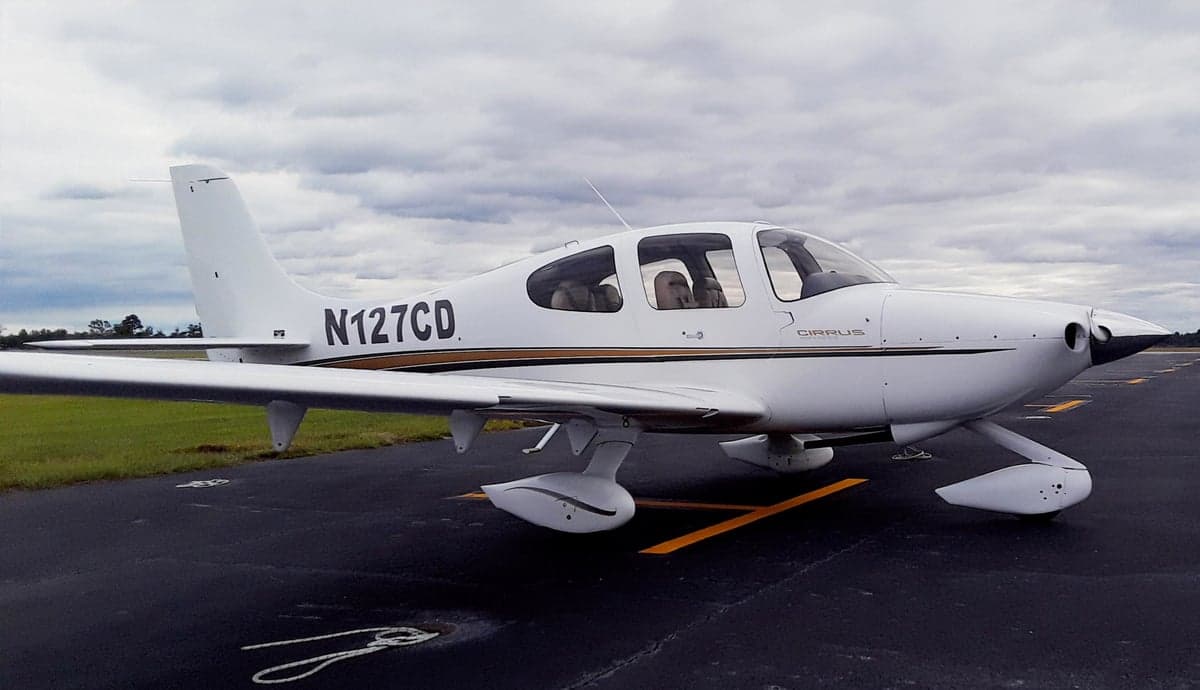 A white Cirrus aircraft with gold and black stripes, registration N127CD, parked on an asphalt ramp under a cloudy sky.