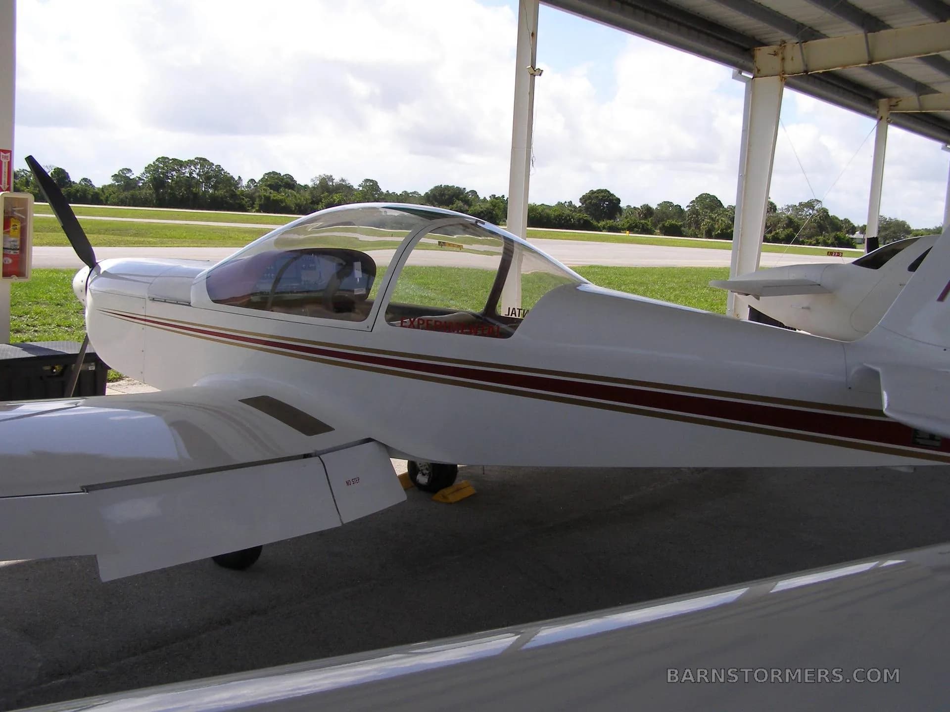 A white experimental aircraft with red and gold stripes, seen from the side under a hangar roof, with a propeller visible.