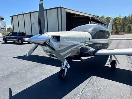A Trinidad GT aircraft is parked on the tarmac in front of a hangar.