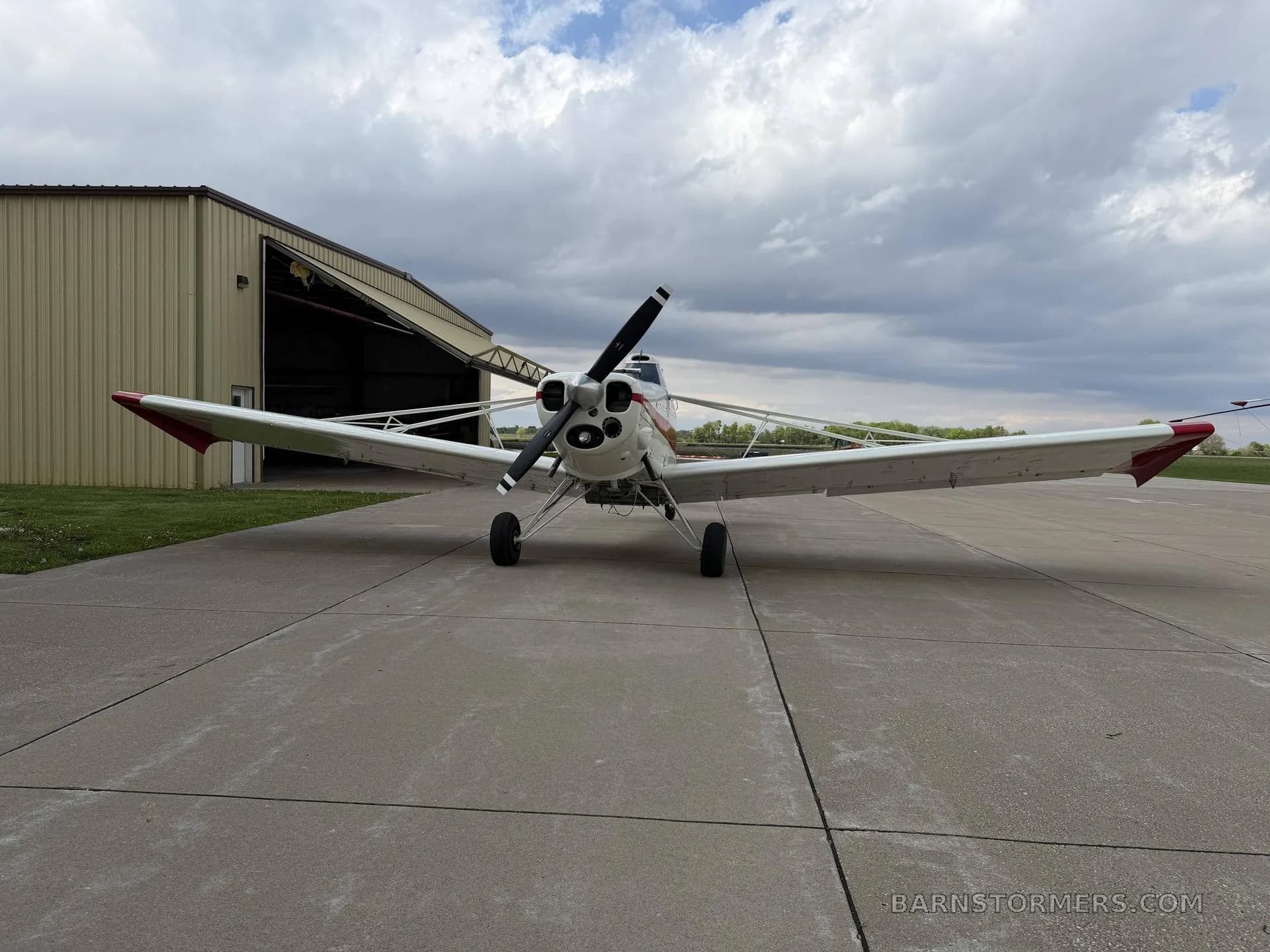 A white and red agricultural aircraft, seen from the front, parked on an airfield in front of an open hangar under a cloudy sky.