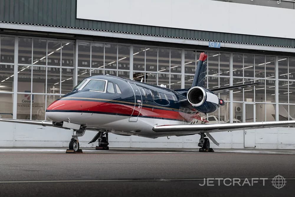 A dark blue, red, and white business jet parked on an asphalt apron, viewed from a front-side angle.