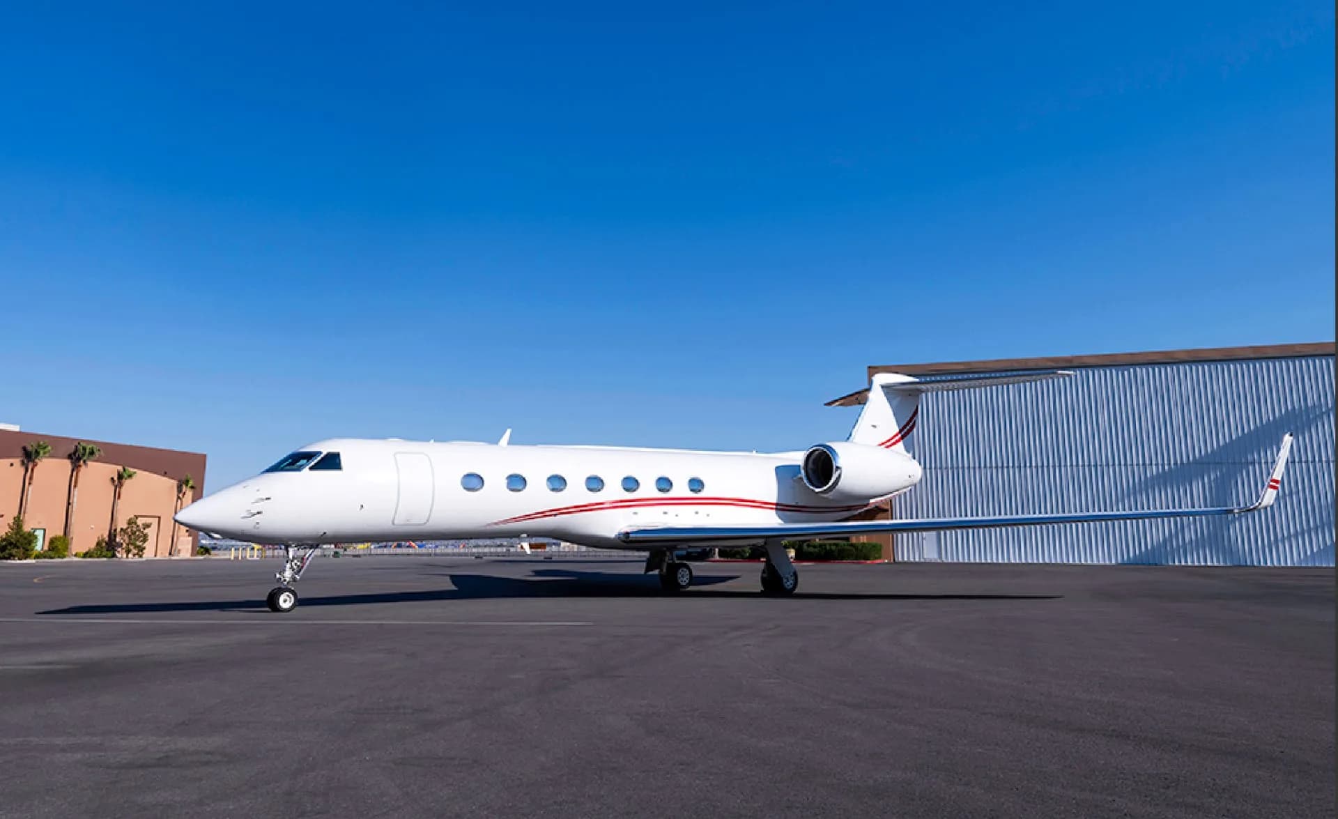 A white Gulfstream private jet with red accent stripes is parked on an airport tarmac in front of a hangar under a clear blue sky.