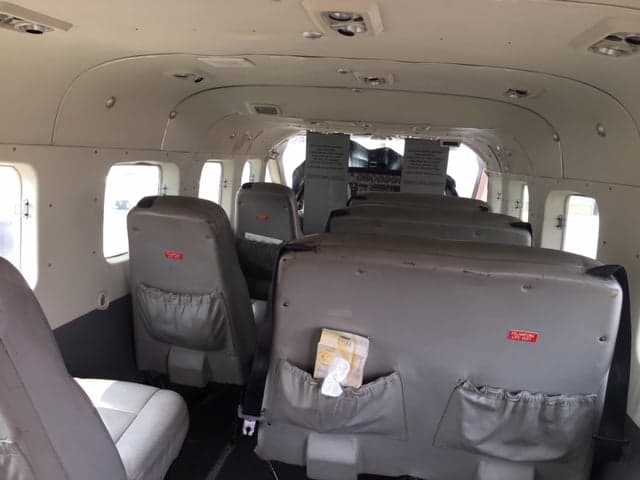 The interior cabin of a Cessna Caravan aircraft, showing grey passenger seats and the view towards the cockpit.