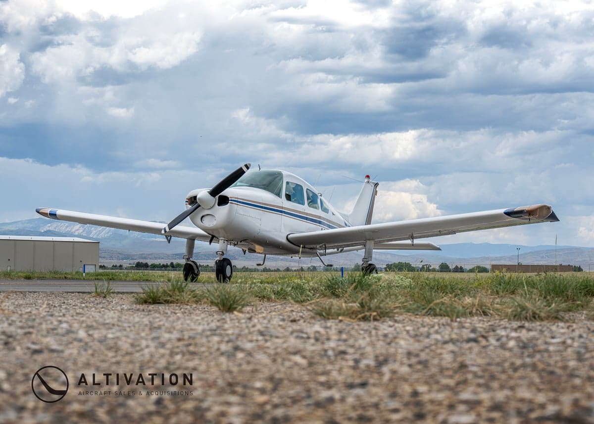 A white and blue Piper aircraft is parked on the tarmac with a cloudy sky in the background.