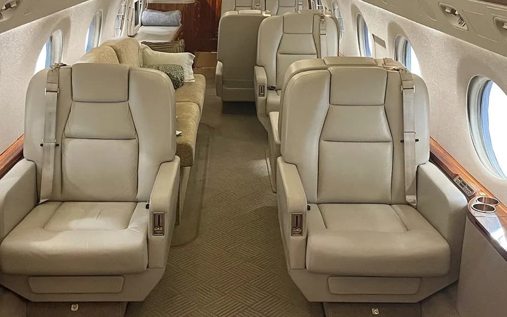 The luxurious cabin interior of a private jet, featuring light beige leather seats arranged in a club configuration and a sofa in the background.