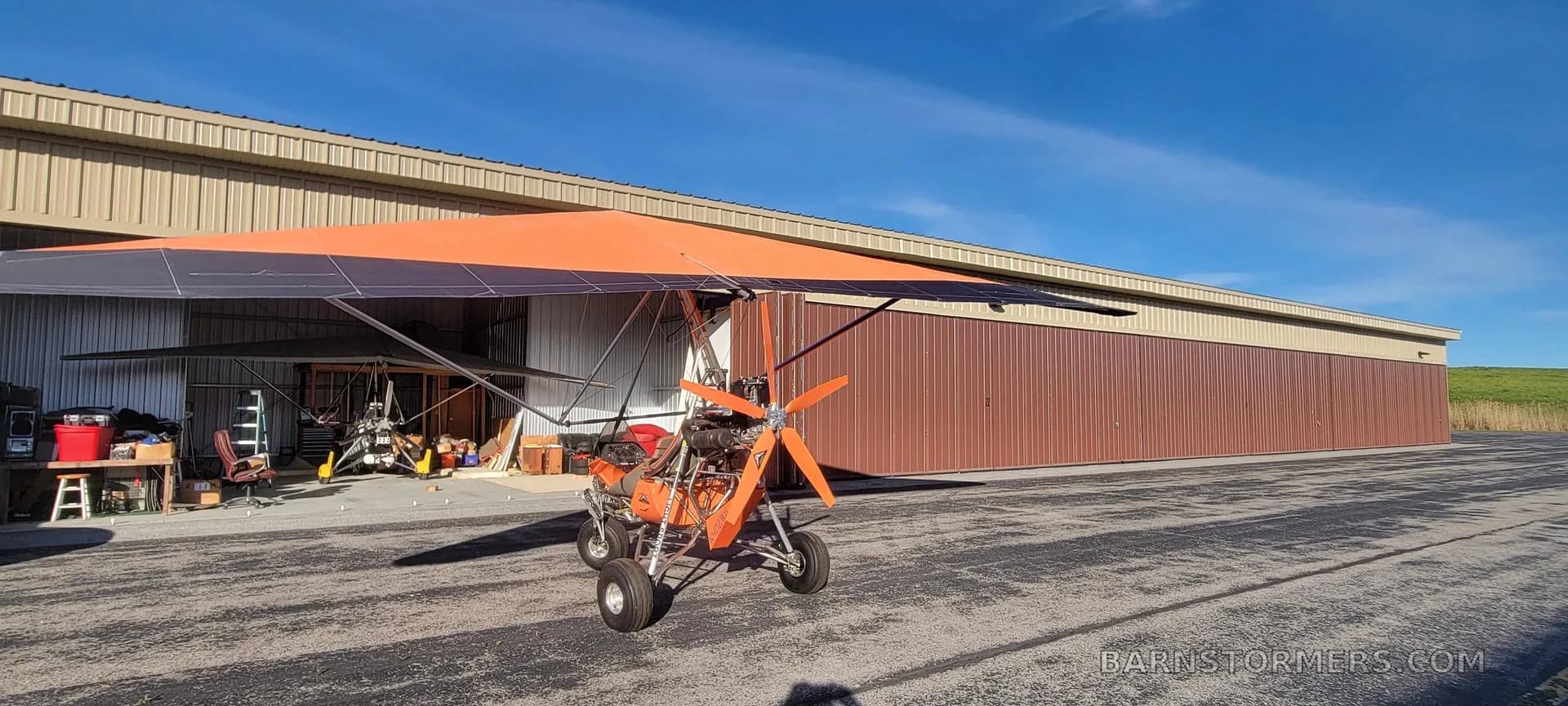 A side view of an orange and black weight-shift control ultralight aircraft parked on an airfield next to a long hangar.