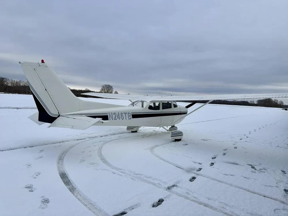 Rear-right view of a white Cessna 172 N246TB with dark blue tail stripes, parked in the snow with tire tracks.