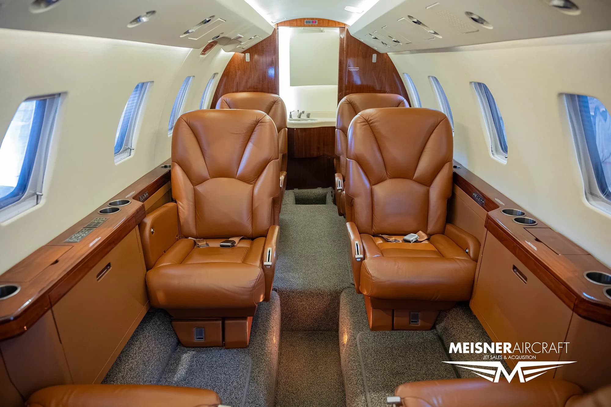 The interior cabin of a private jet with brown leather seats, looking towards the lavatory area at the rear.
