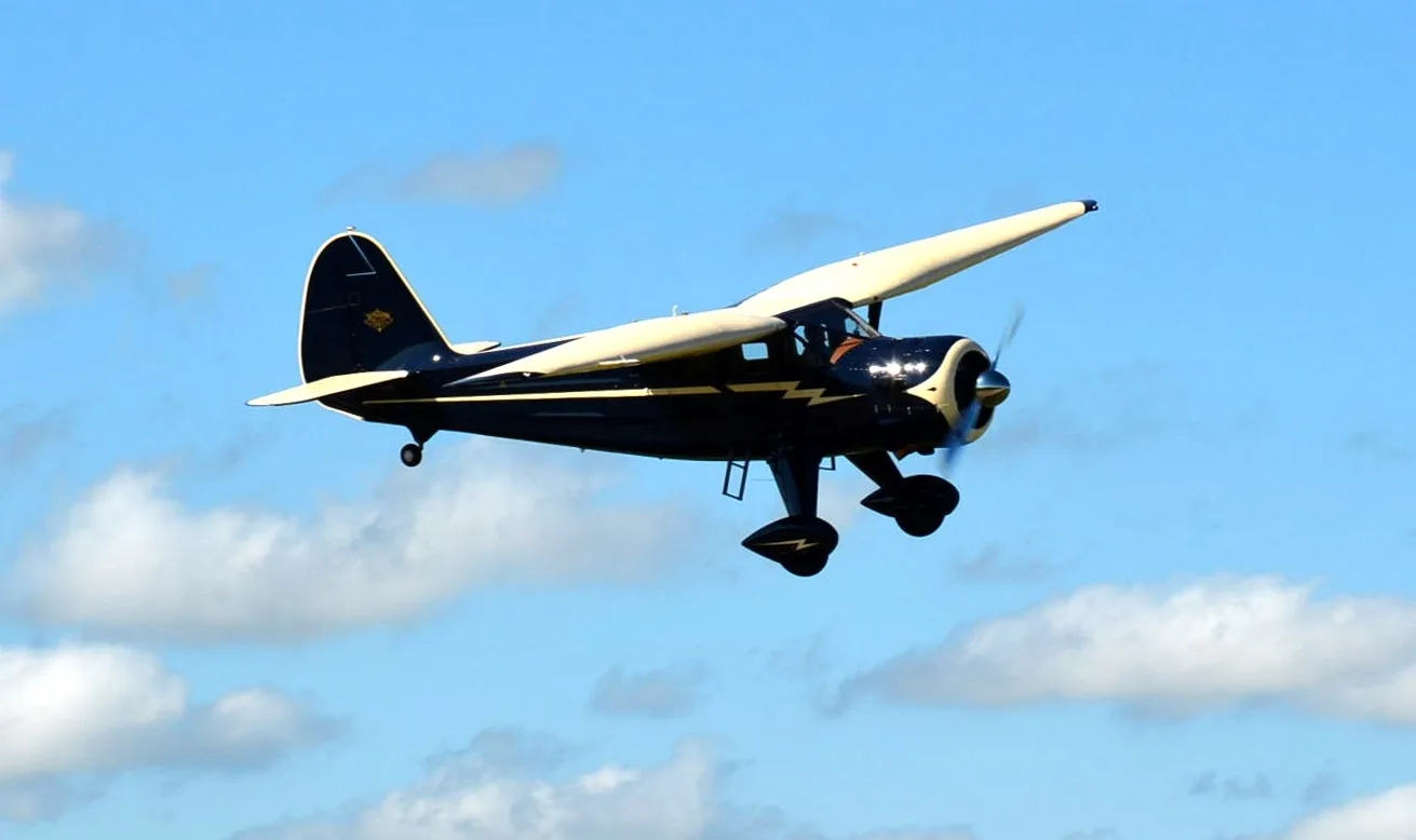 A dark blue and cream vintage aircraft with a lightning bolt design is flying against a partly cloudy blue sky.