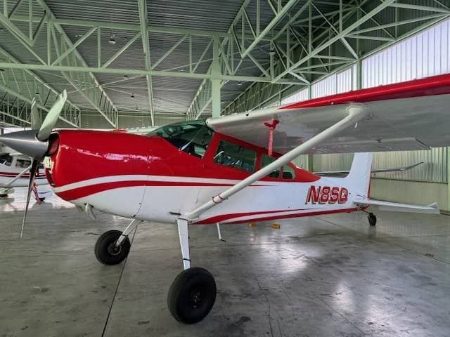 A red and white Cessna 180/185 aircraft, tail number N8SQ, parked inside a hangar.