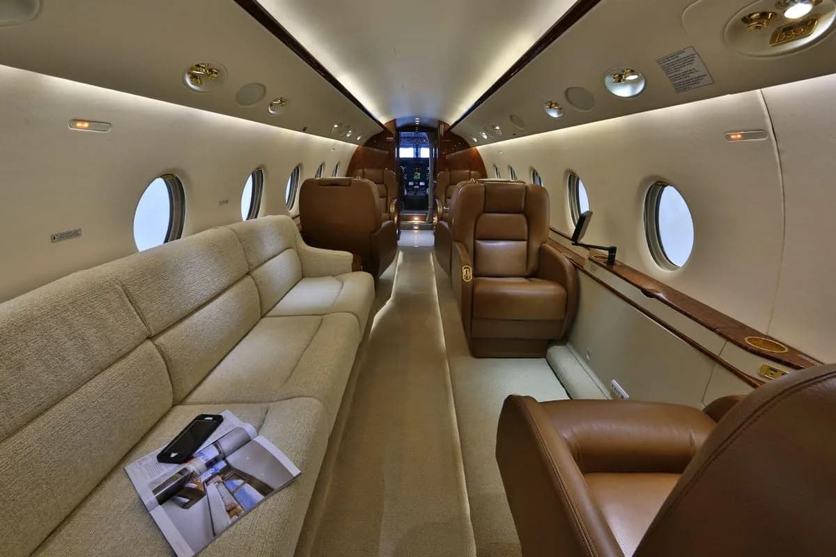 The spacious cabin of a business jet features a long beige sofa, brown leather seats, and oval windows.