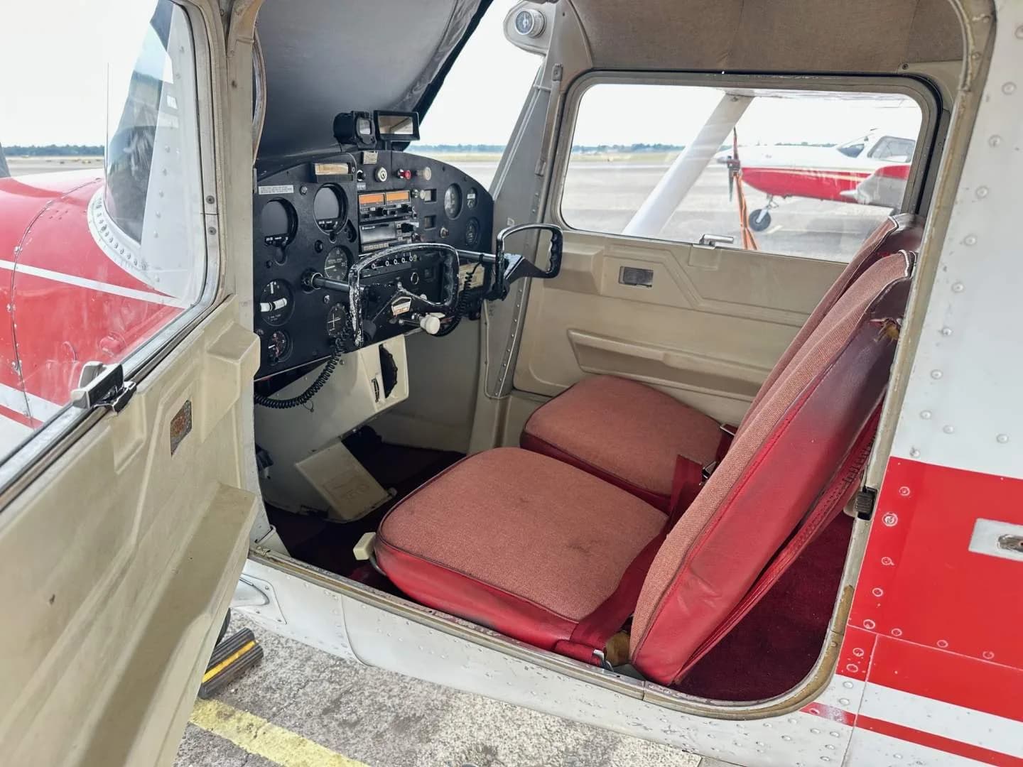 The interior of the Cessna 150, showing the red seats and the instrument panel from the open door.