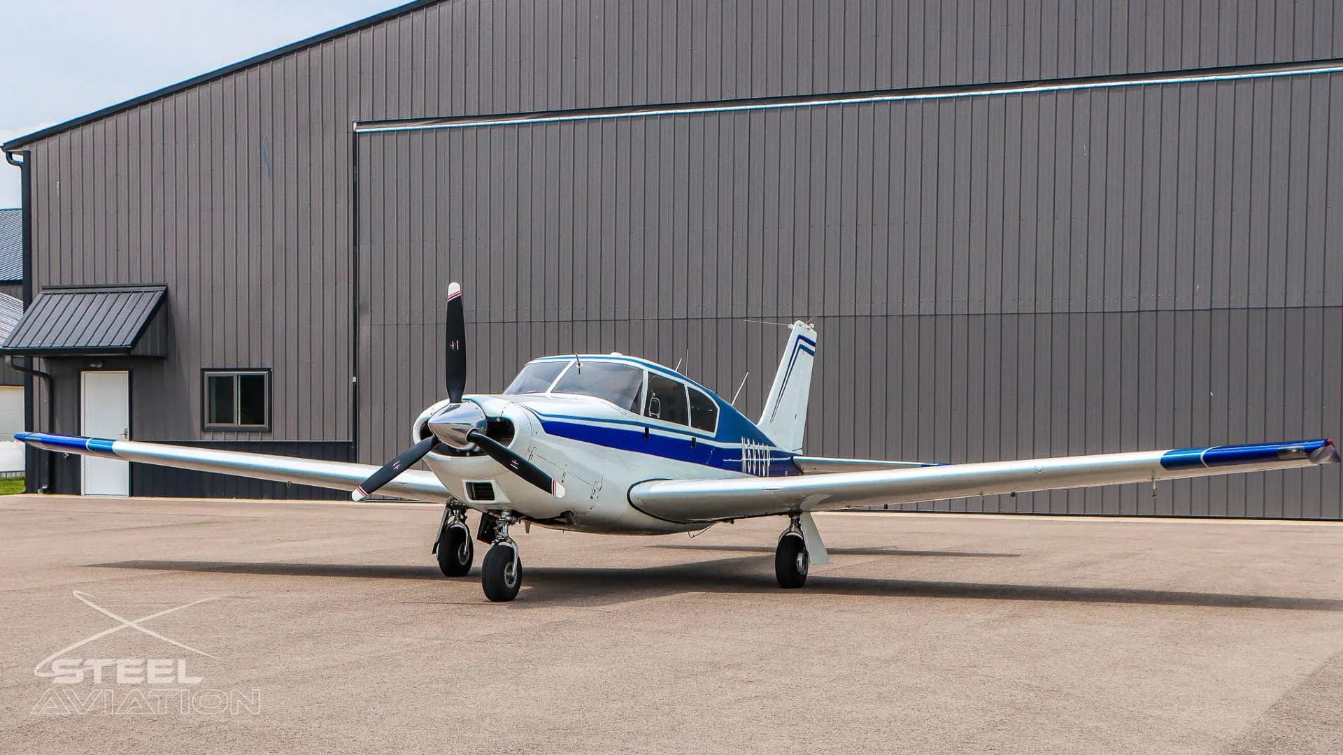 A front three-quarter view of a white and blue single-engine aircraft parked in front of a dark gray hangar.